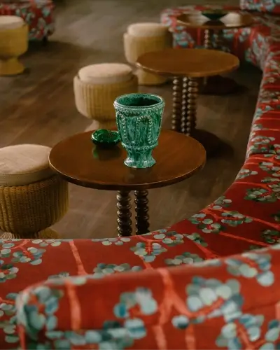 Wooden round table with a decorative green goblet and small green dish on top, surrounded by wicker stools and a red floral patterned sofa in a cozy indoor seating area.