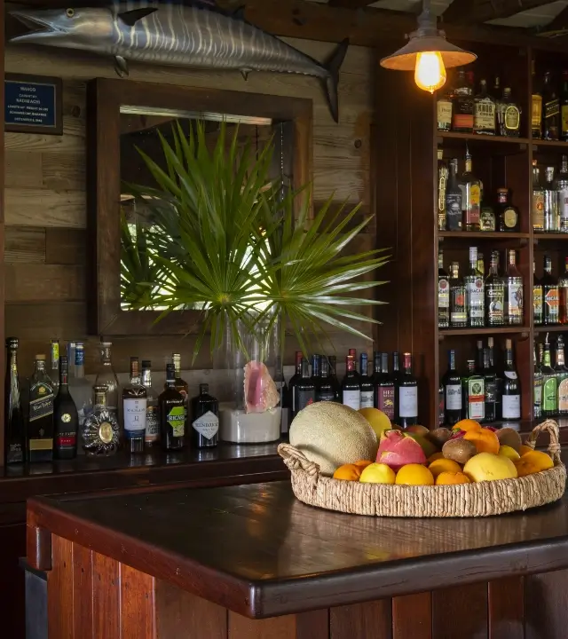 Wooden bar interior with shelves of liquor bottles, a large mounted fish above a mirror, a glass vase holding green palm leaves, and a basket of assorted fruits on the bar counter.