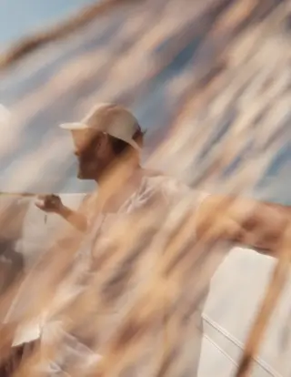 Bearded man wearing a backward baseball cap and light clothing, standing on a boat with hands on the railing, viewed through blurred tall grass or reeds.