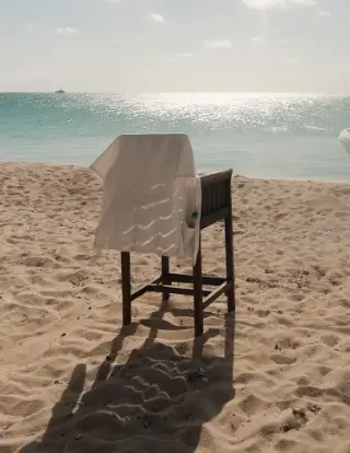 Wooden high chair covered with a white cloth standing on sandy beach with ocean and sparkling water in the background under partly cloudy sky.