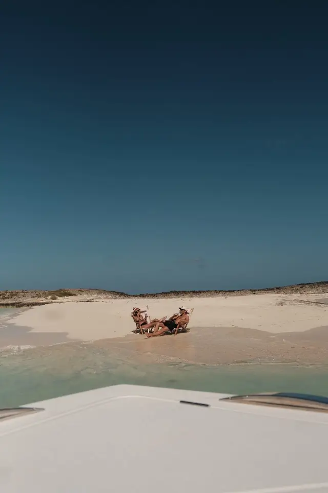 Two people lounging on beach chairs on a sandy shore near clear, shallow water, viewed from a boat.