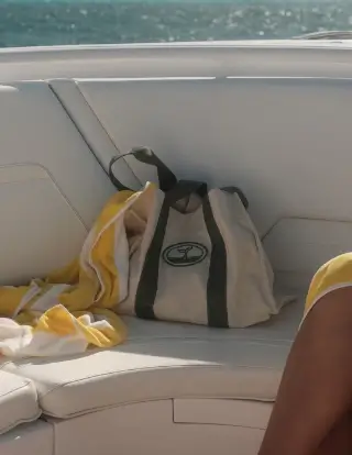Beige and black tote bag placed on a white cushioned seat on a boat with a yellow and white striped towel beside it, ocean visible in the background.