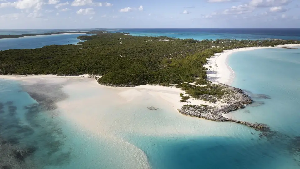 Aerial view of a tropical island with dense green vegetation and surrounding white sandy beaches bordered by clear blue ocean waters under a partly cloudy sky.