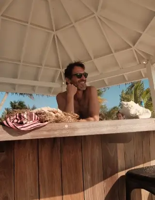 Smiling man wearing sunglasses leaning on a wooden bar under a white canopy with a blue sky, palm trees, and a large seashell in the background.