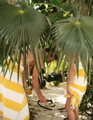 Two people wrapped in yellow and white striped towels with large green palm leaves partially obscuring them, standing outdoors on sandy ground with greenery in the background.