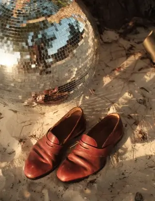Brown leather dress shoes placed on sandy ground near a large silver disco ball reflecting light and surroundings.