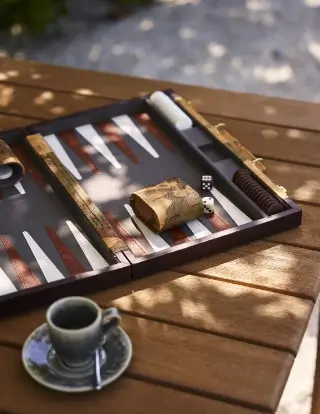 Open backgammon board with dice and doubling cube on wooden table, next to a cup of coffee on a saucer with a spoon.
