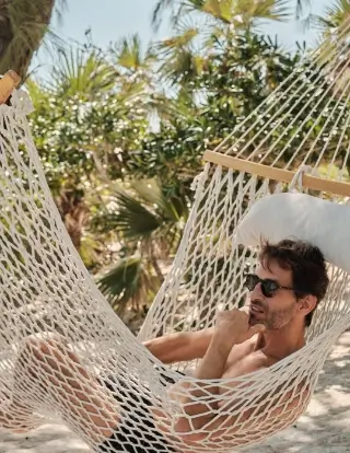A man wearing sunglasses relaxing on a white hammock outdoors with palm trees in the background.