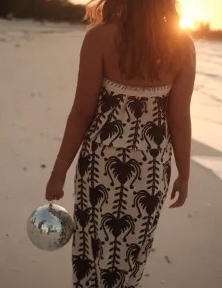 Woman walking away on a sandy beach at sunset, wearing a sleeveless dress with a dark tropical leaf pattern and holding a silver disco ball handbag in her right hand.