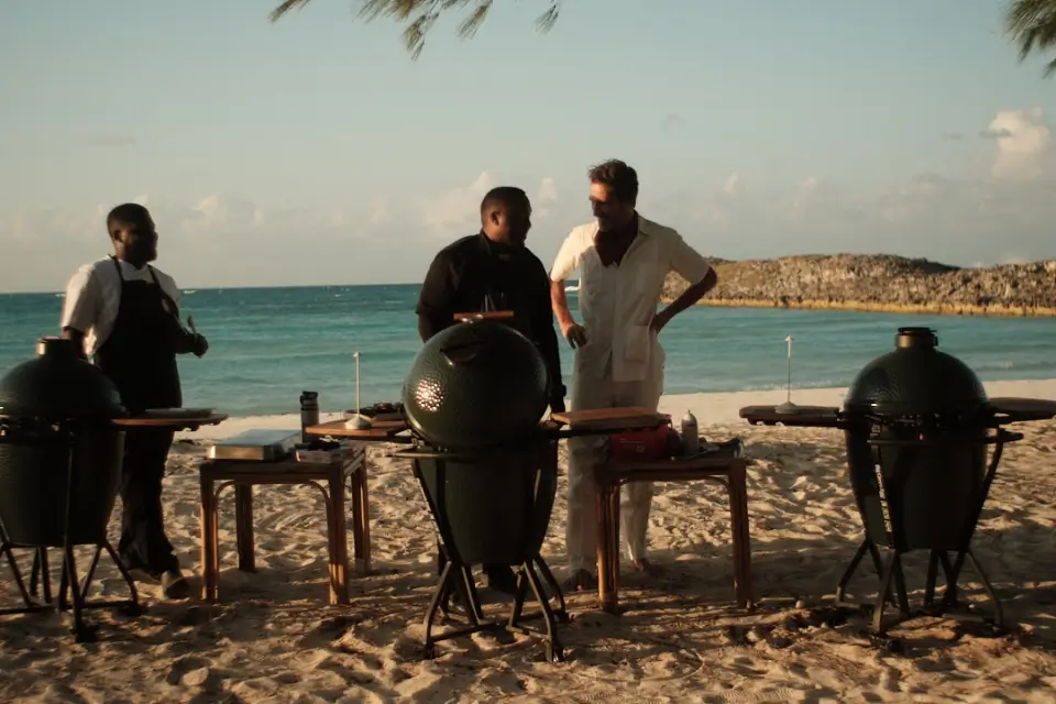 Three men standing on a sandy beach near the ocean, with three green kamado-style grills and wooden tables set up around them under a partly cloudy sky.