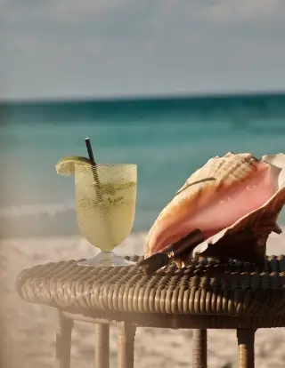 A cold lemon drink with a straw and a lemon wedge on the rim sits on a round wicker table next to a large pink seashell on a sandy beach with ocean water in the background.