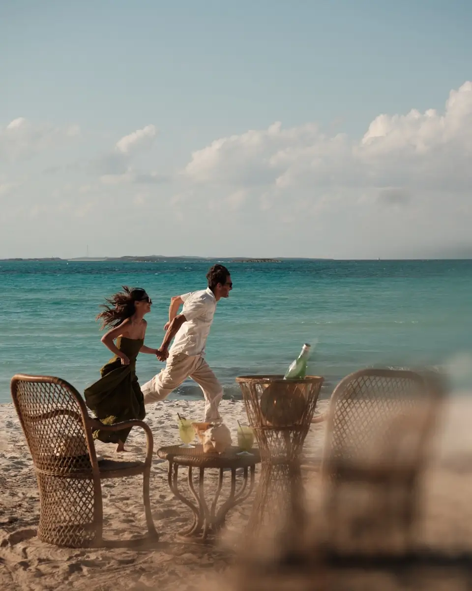 A man and woman holding hands and running on a sandy beach near blue ocean water with wicker chairs and a table with drinks in the foreground.