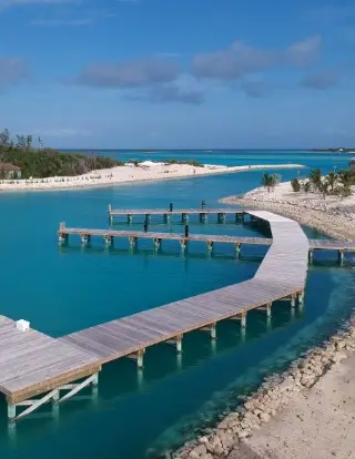 Wooden curved dock extending over clear turquoise water of a marina with sandy shores and sparse vegetation under a blue sky with some clouds.