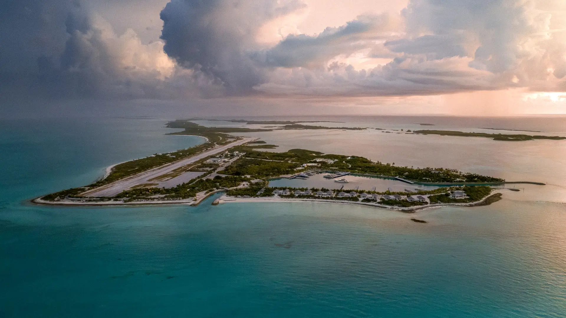 Aerial view of a narrow island with a marina featuring boats docked in a protected harbor, surrounded by turquoise waters and a cloudy sky at sunset.