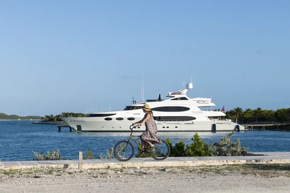 Person wearing a hat and dress riding a bicycle along a waterfront with a large white yacht docked in the background under a clear blue sky.