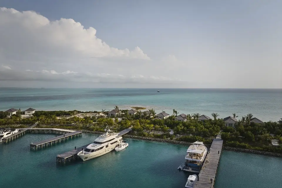 A serene marina with two yachts docked at wooden piers, lush greenery and small cottages behind, and a calm blue ocean under a partly cloudy sky.