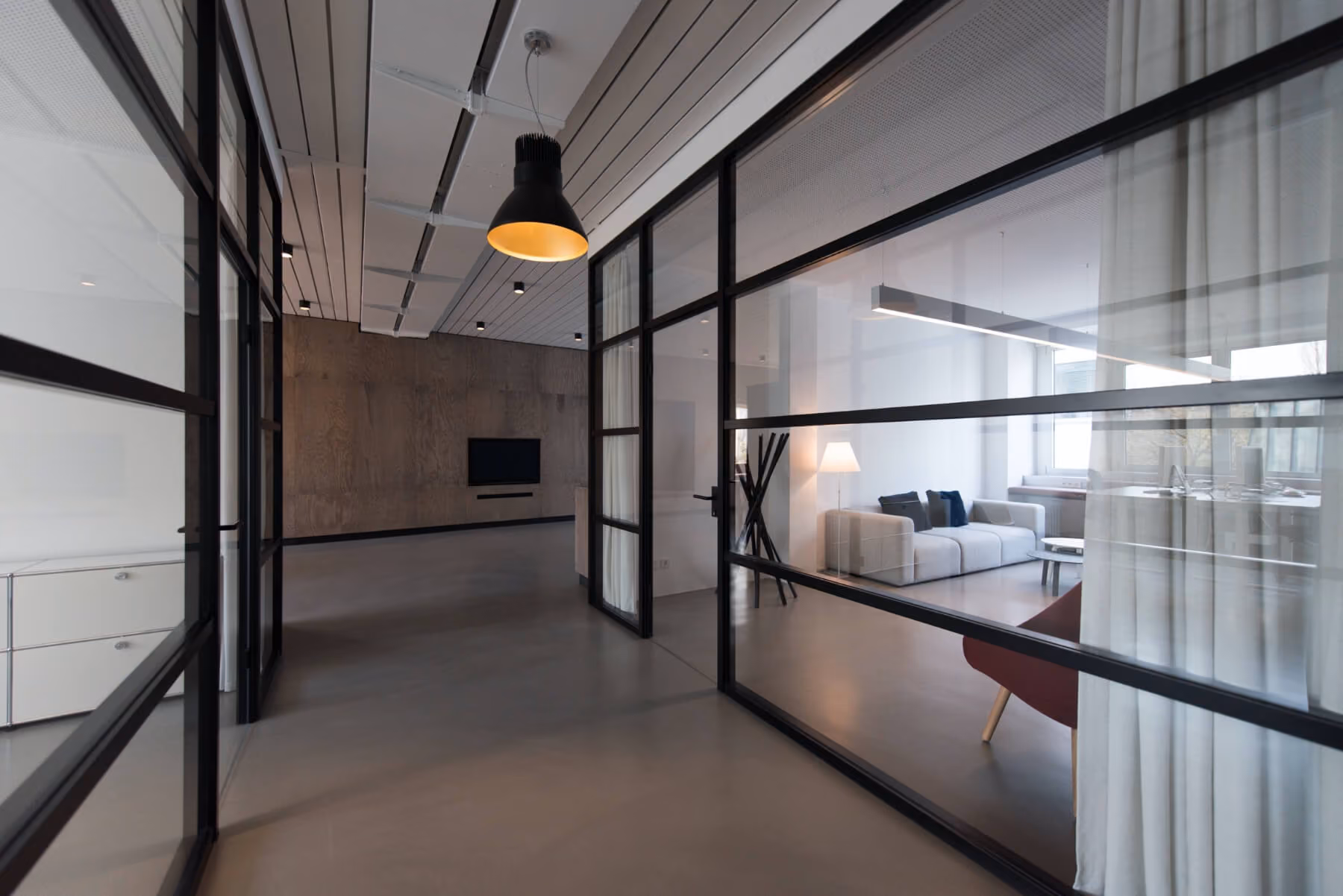 Modern office hallway with glass walls revealing a white sofa and lamp in a meeting room.