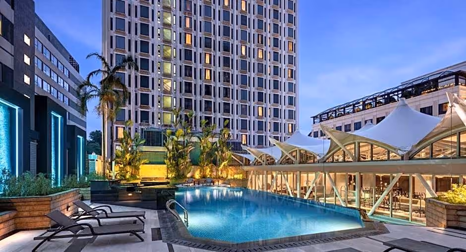 Outdoor view of Peninsula Excelsior hotel pool area with lounge chairs, palm trees, and a tall building in the background at dusk.