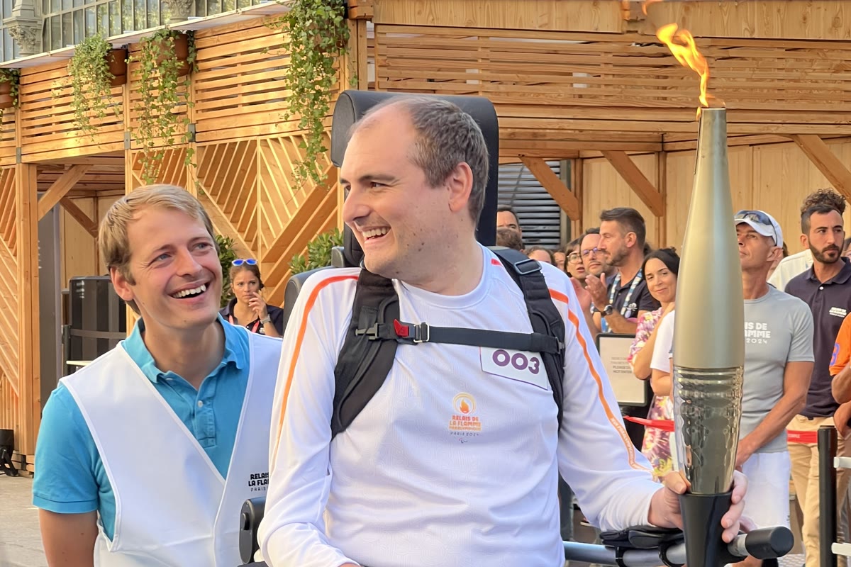 Two men at the 2024 Paralympic torch relay in Paris, one walking in a Wandercraft exoskeleton and carrying the torch