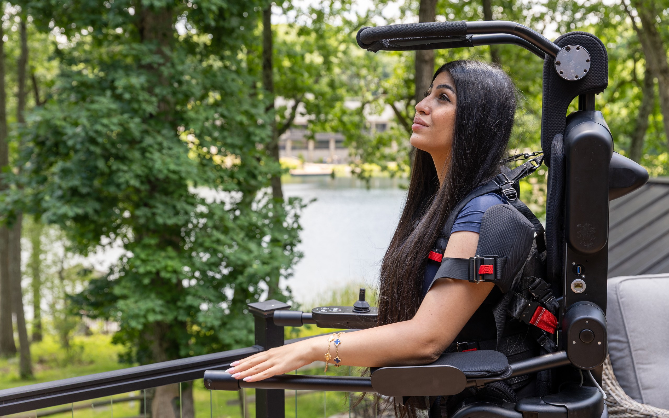 A woman upright in the Eve personal exoskeleton on a lakeside deck, one hand resting on the railing