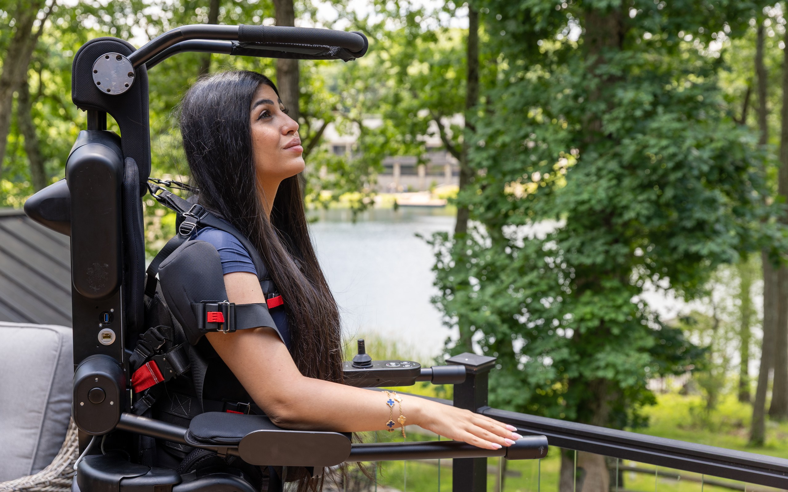 A woman upright in the Eve personal exoskeleton on a lakeside deck, one hand resting on the railing