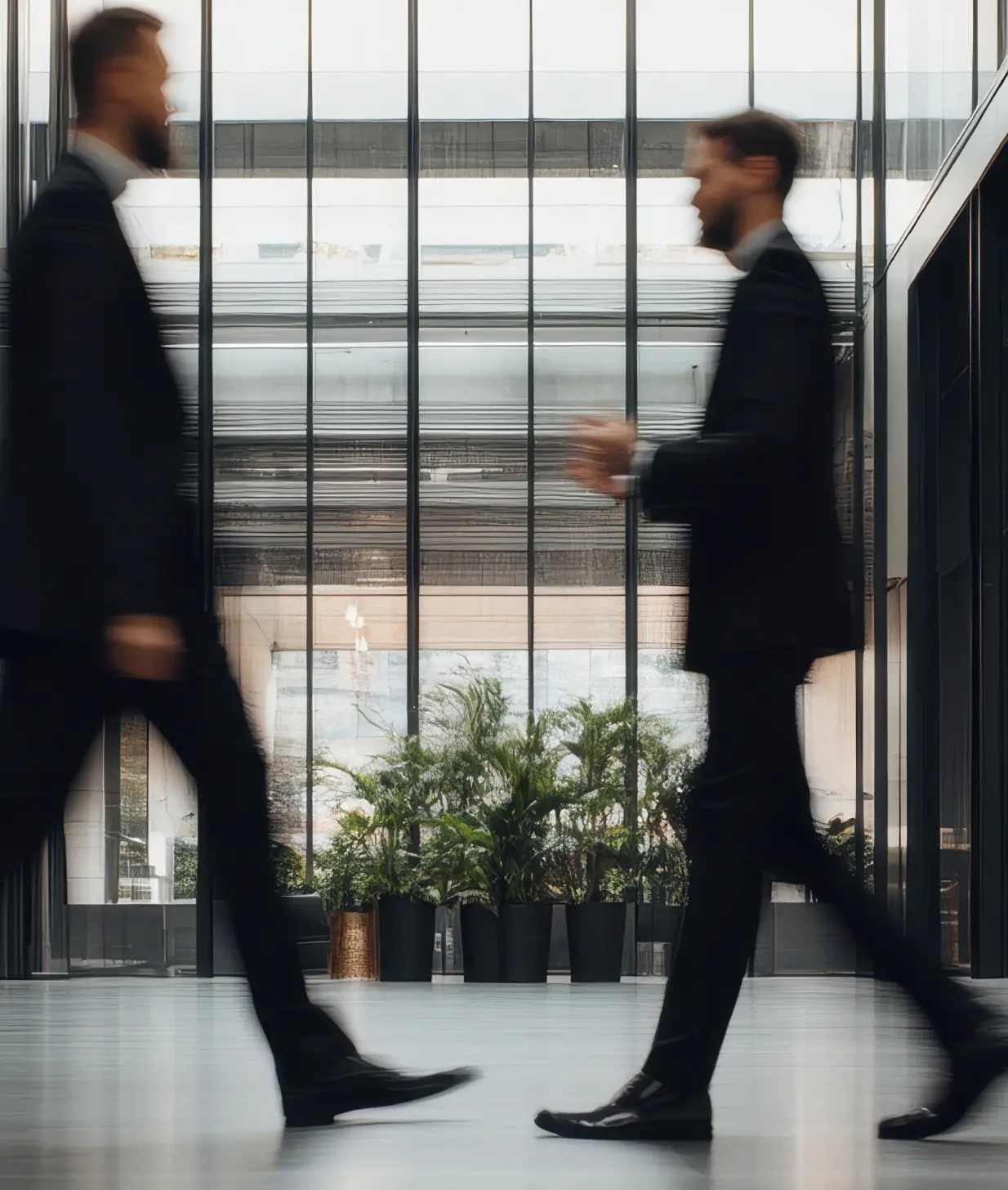 Two men in black suits walking past each other inside a modern building lobby with large glass windows and several large potted plants.