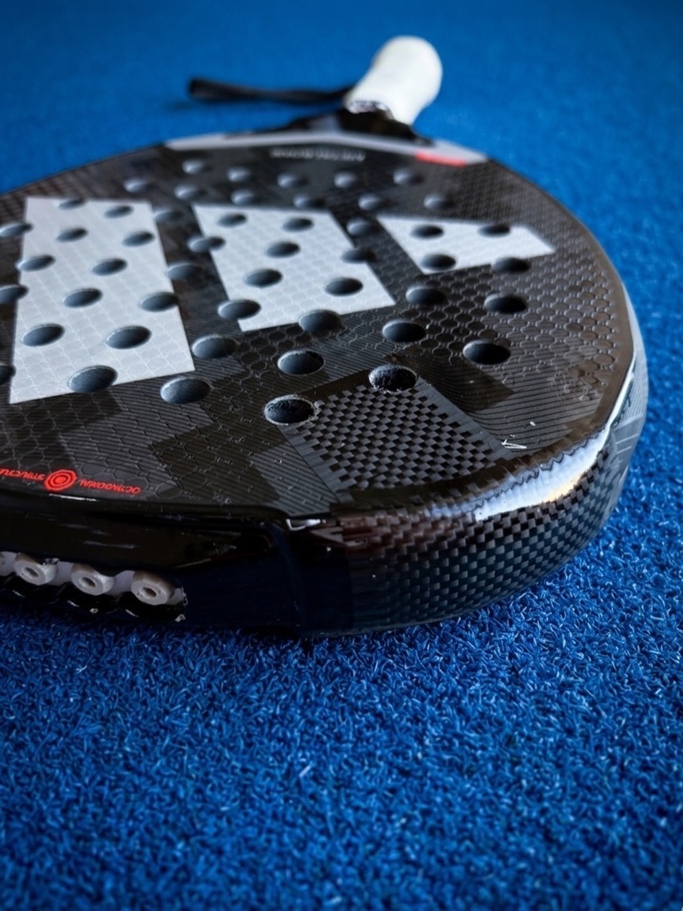 Close-up van het blad van een zwart-wit padelracket met stervormig gaatjespatroon op de padelbaan