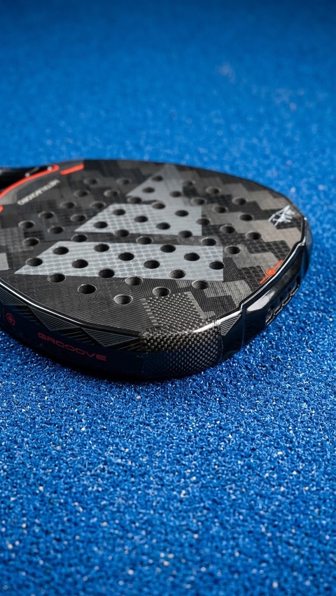 Close-up van een zwart padelracket met camouflagepatroon en oranje accenten op de padelbaan