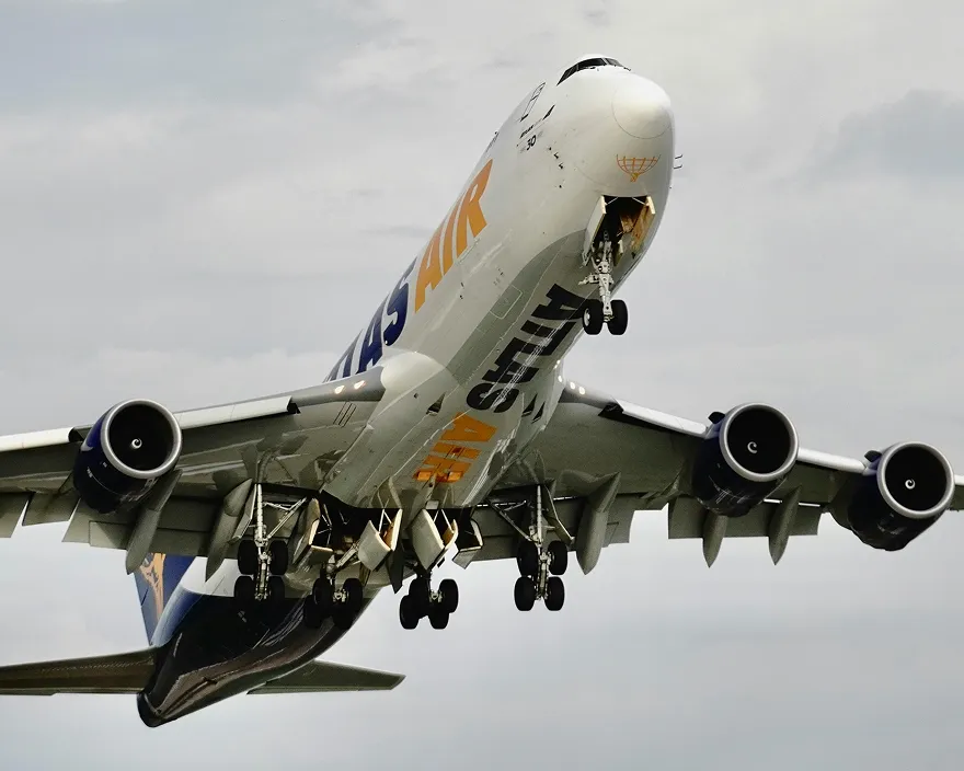 Cargo airplane with open nose gear doors flying against a cloudy sky.
