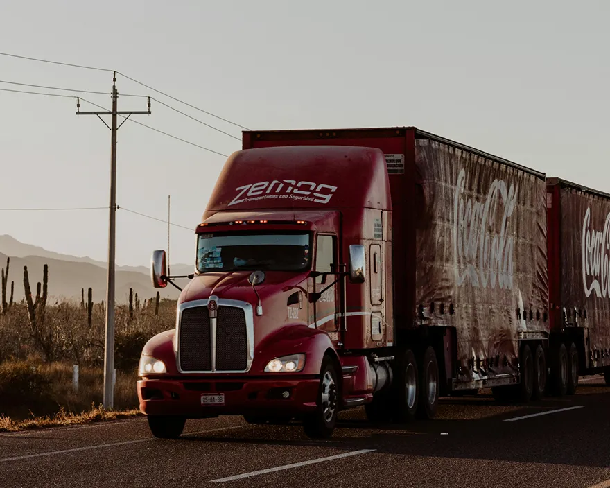 Red semi-truck with Coca-Cola trailers driving on a road at sunset in a desert area.
