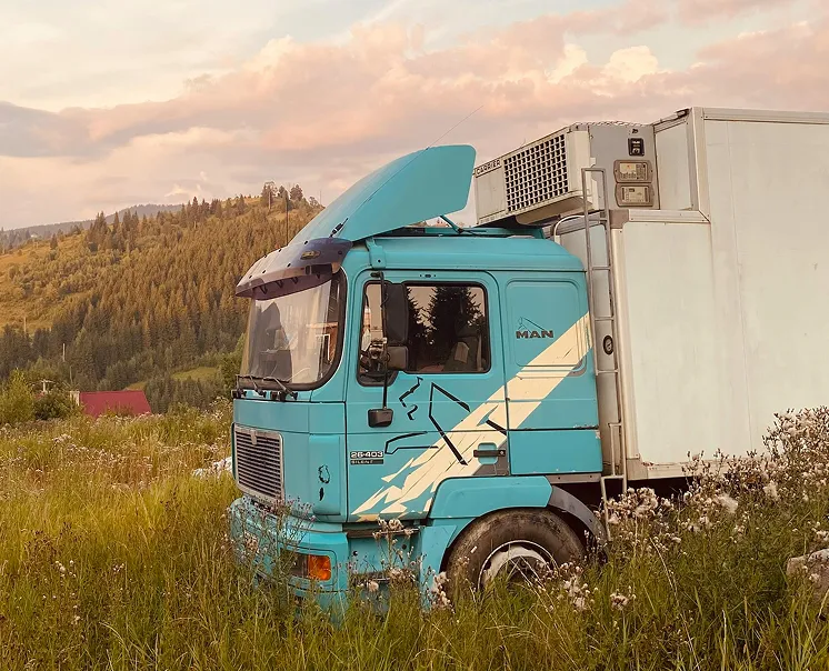 Blue MAN truck with white design parked in tall grass near forested hills at sunset.