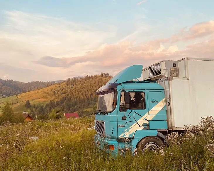 A blue truck with an open hood in a grassy field with hills and houses in the background at sunset.