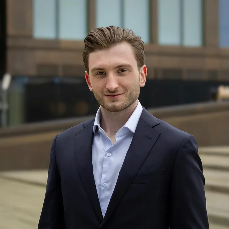 Young man with light brown hair in a dark suit jacket and light blue shirt stands outdoors, smiling slightly.