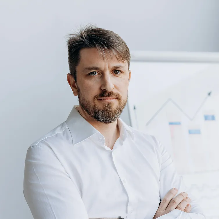 Serious man with beard and crossed arms wearing a white shirt in front of a blurred chart.