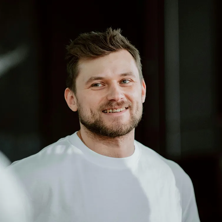 Smiling man with short brown hair and beard wearing a white shirt against a dark background.