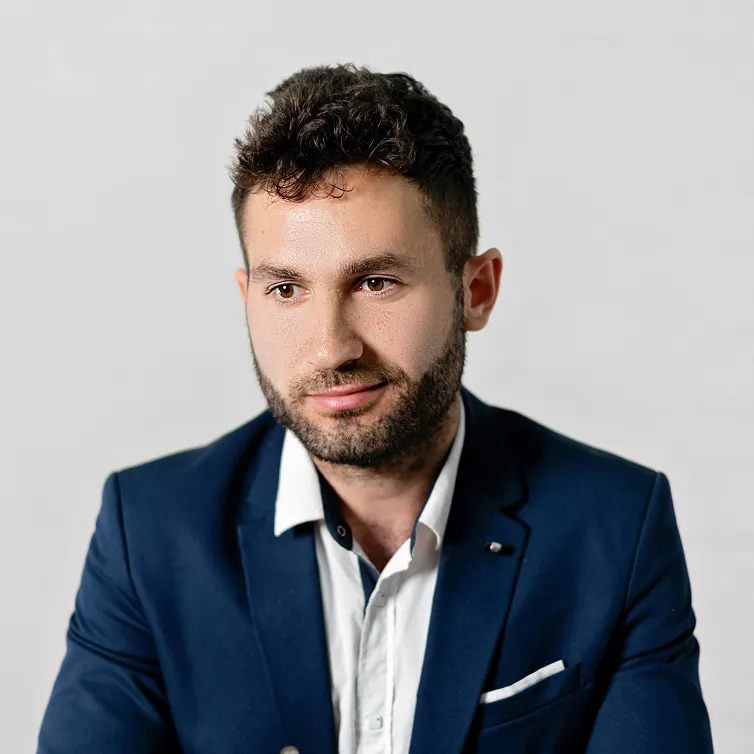 Young man with short curly hair and beard wearing a blue blazer and white shirt on gray background.
