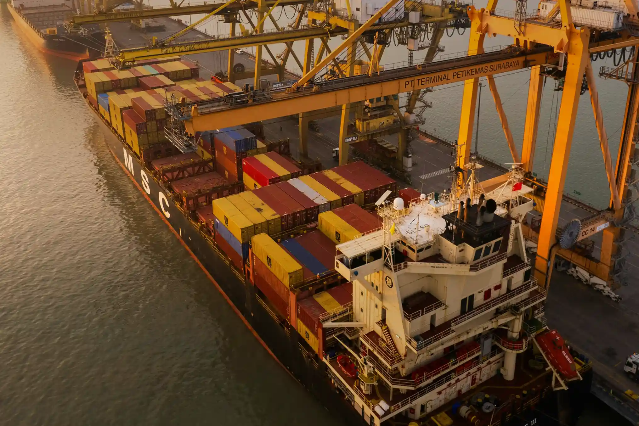 A cargo ship loaded with colorful containers docked at a port terminal with large yellow cranes.