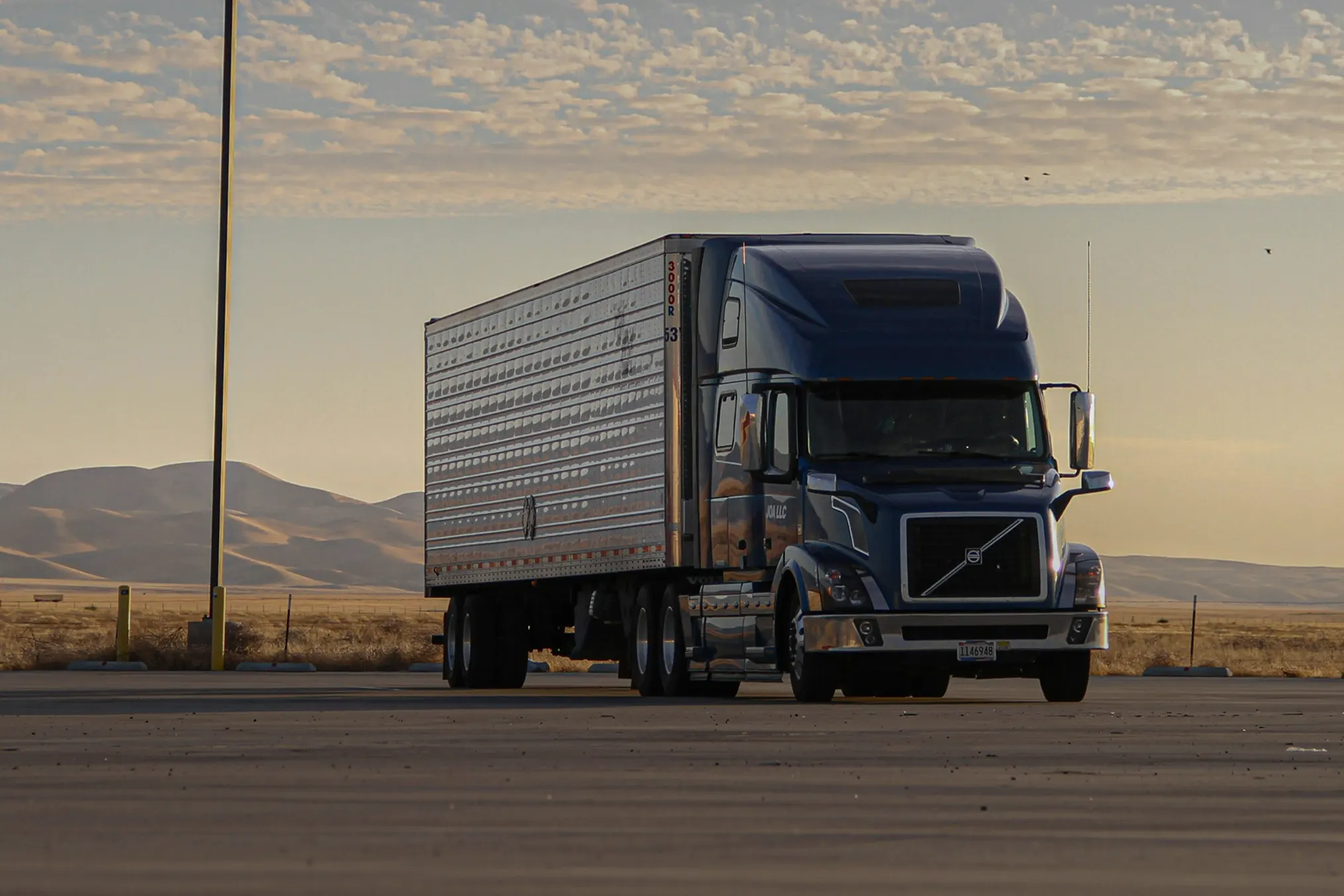 Blue semi-truck with a large silver trailer parked on a flat surface at sunset with hills in background.