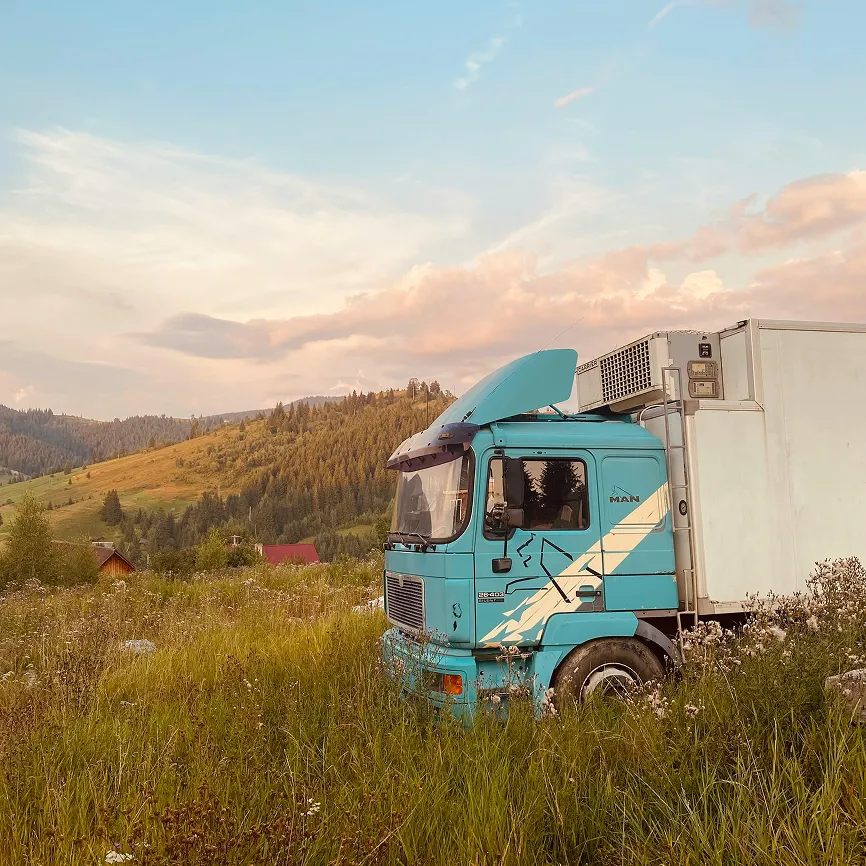 Blue truck with raised hood parked in grassy field with hills and trees in background under a cloudy sky.
