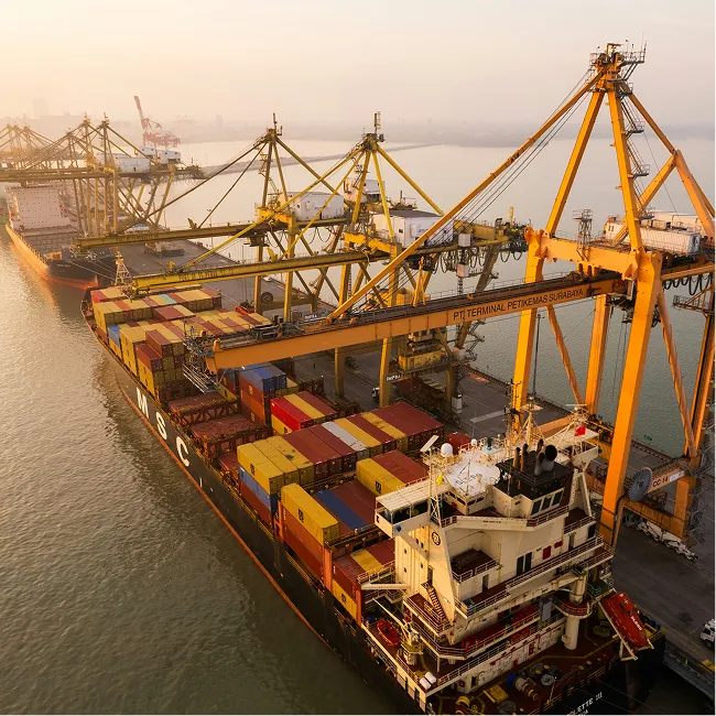Large cargo ship loaded with colorful containers docked at port with yellow cranes during sunset.