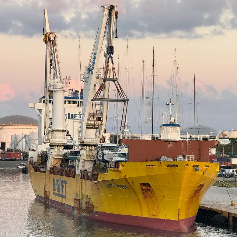 Large yellow cargo ship named Happy River docked at port with cranes and sailboats behind it.