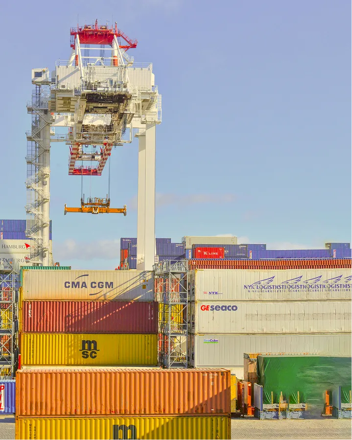 Port crane lifting a container amidst stacked shipping containers under clear sky.