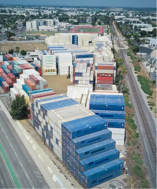 Aerial view of stacked shipping containers near a railway and road in an industrial area.