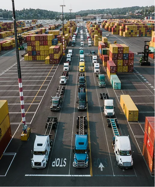 Overhead view of semi trucks lined up in lanes at a shipping container port with stacks of containers.