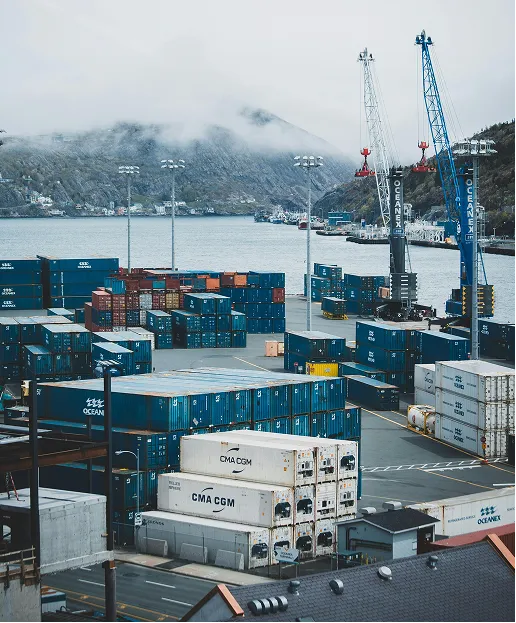 Shipping containers stacked at a port with cranes and fog-covered mountains in the background.