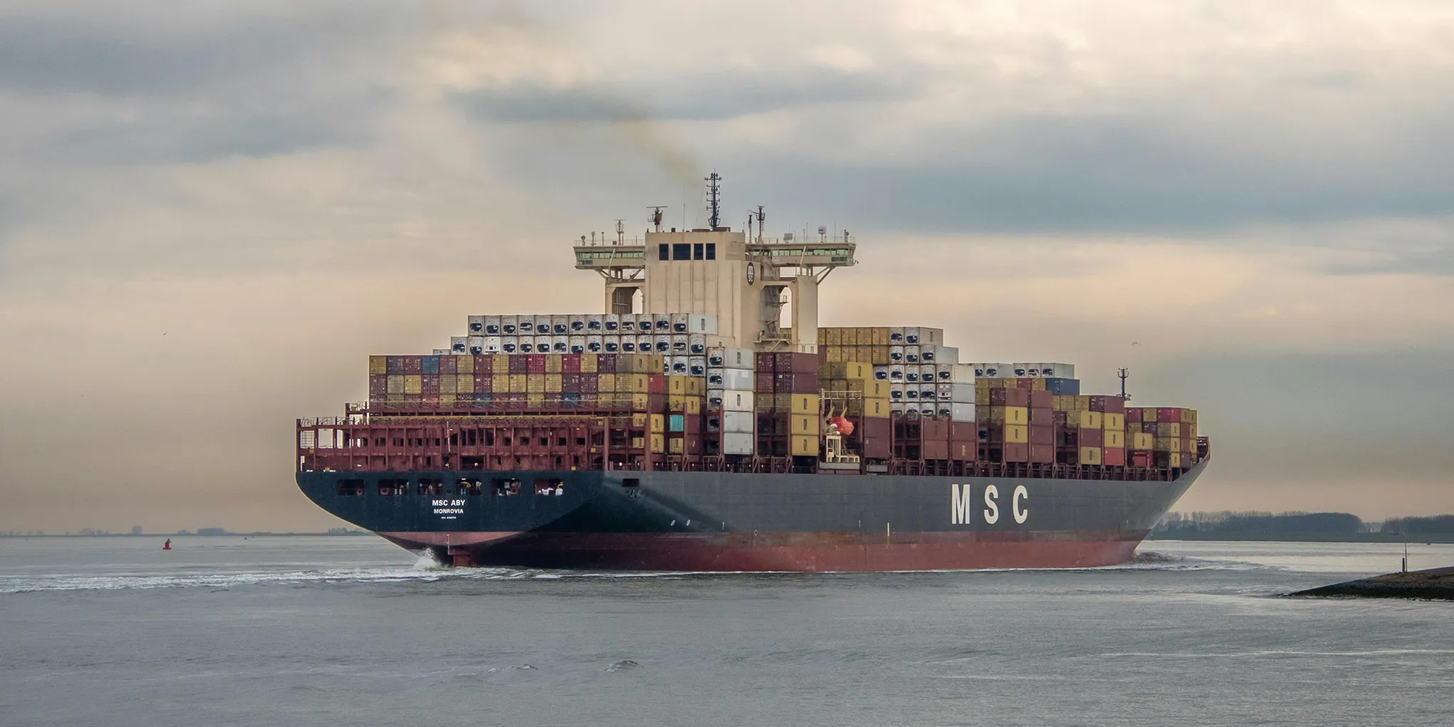 Cargo ship MSC ABY Monrovia loaded with shipping containers sailing on calm water under cloudy sky.