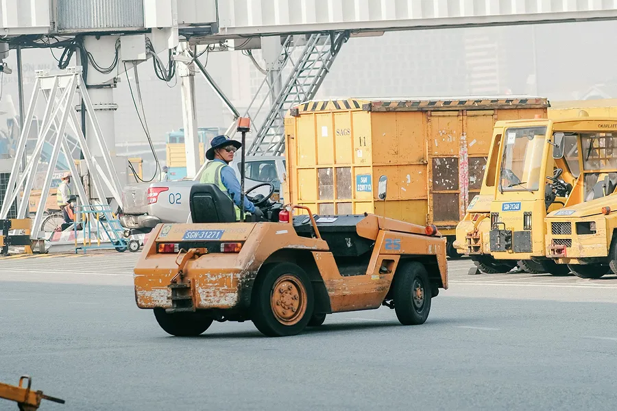 Worker driving an orange utility vehicle on industrial site with yellow equipment in the background.
