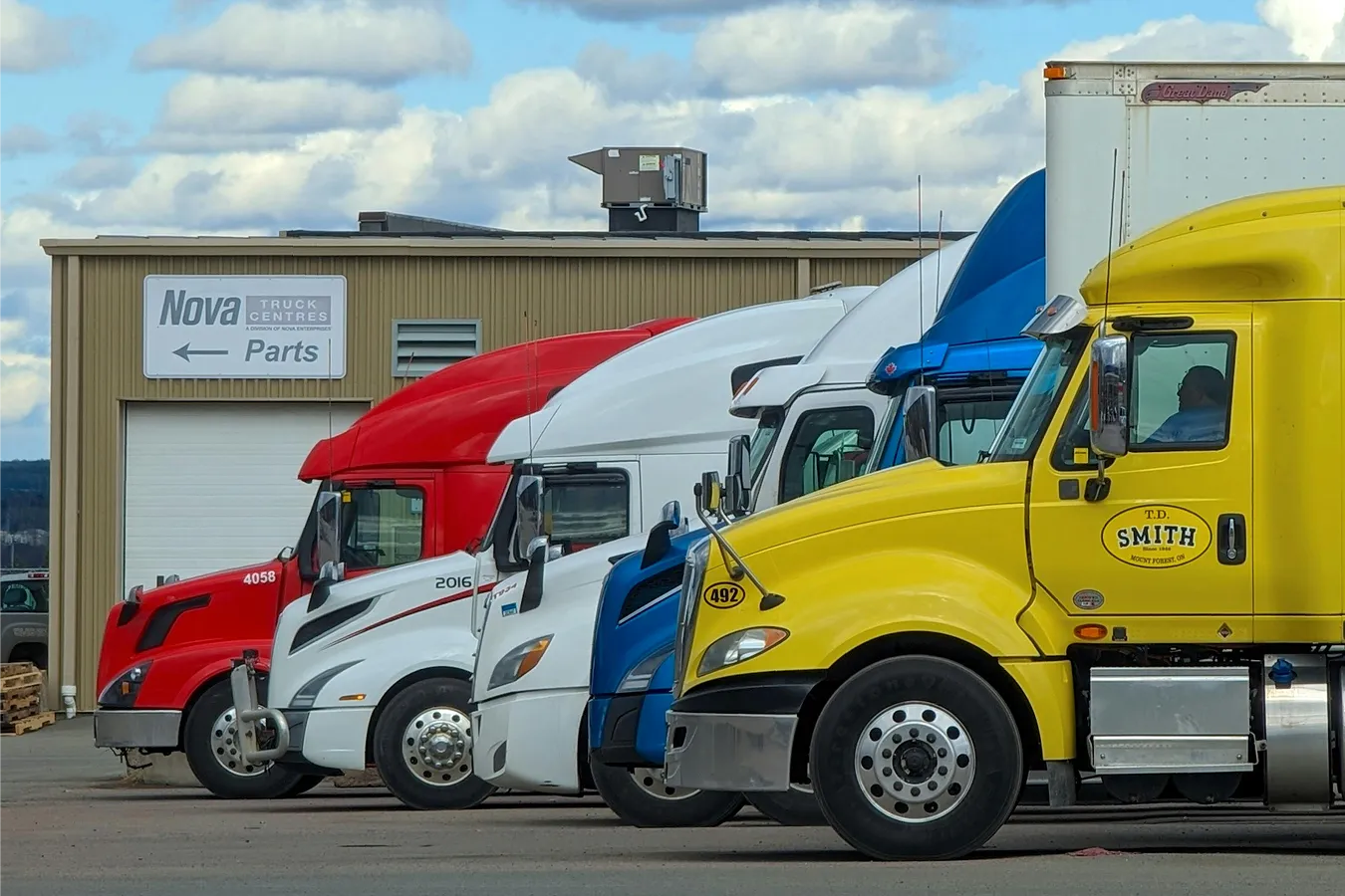 Side view of five parked semi trucks in red, white, blue, and yellow near a parts center building.