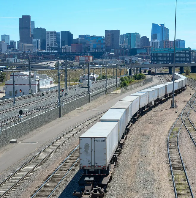 Long freight train carrying white shipping containers on curved tracks near an urban city skyline.