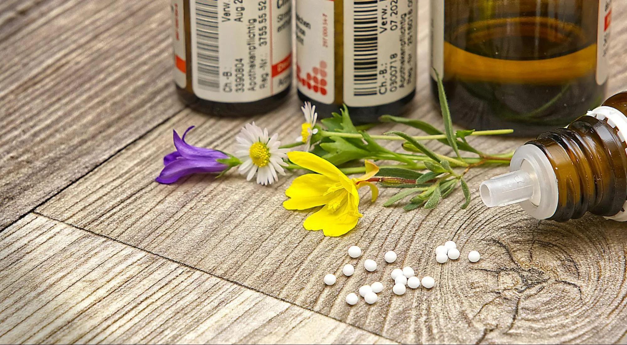 Small white homeopathic pellets and dropper bottles lie on a wooden surface beside colorful wildflowers.